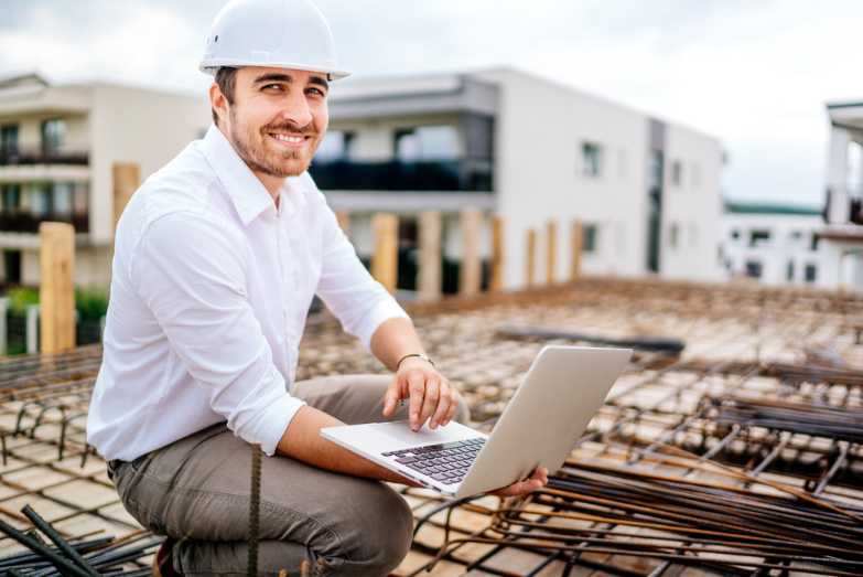 Bauleiter sitzt mit Notebook auf dem Rohbau eines Gebäudes.